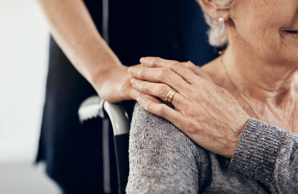 A caregiver’s hand resting gently on the shoulder of an elderly woman in a wheelchair